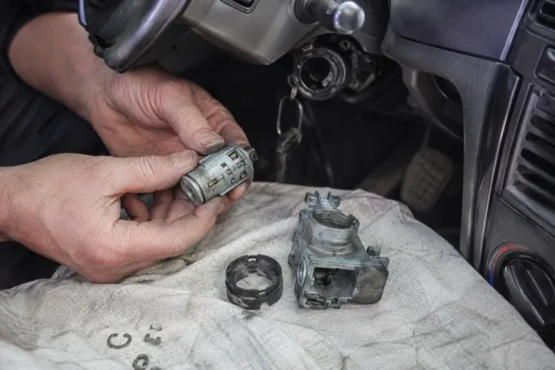 Hands holding a removed ignition cylinder beside a car steering column, with the housing and small parts on a cloth. — Ignition Lock Repair, Wasaga Beach